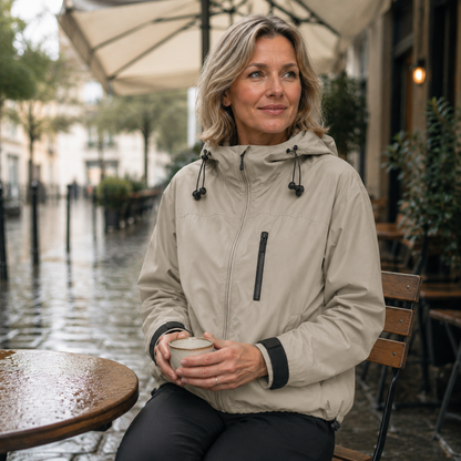 Beige korte damesjas met capuchon, gedragen door een zittend model op een terras.
