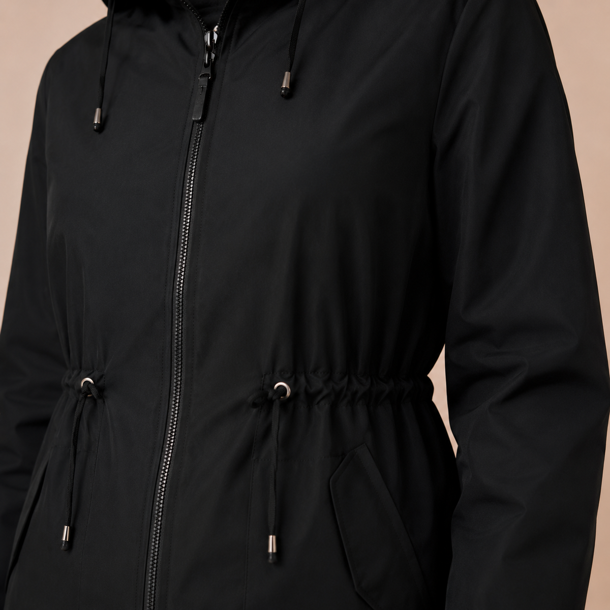 Regenjas dames lang – waterafstotende parka met capuchon en taillekoord in zwart – detailopname op beige studioachtergrond