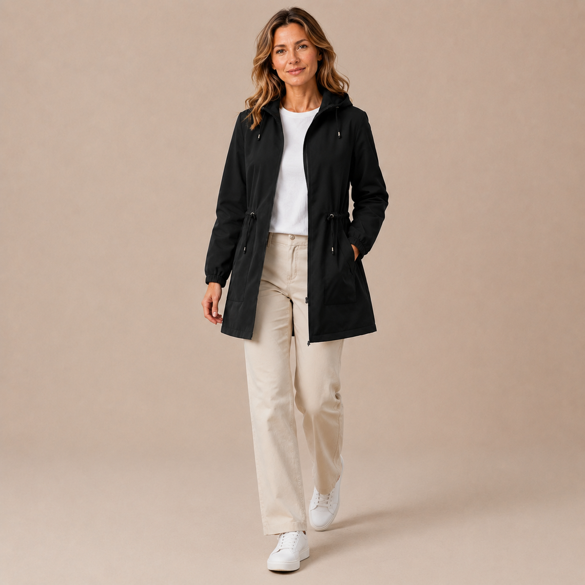 Regenjas dames lang – waterafstotende parka met capuchon en taillekoord in zwart – lifestylebeeld op beige studioachtergrond